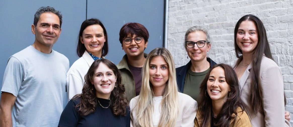 The Reservoir team: eight people, smiling, standing and sitting together in front of a grey door and a white brick wall.